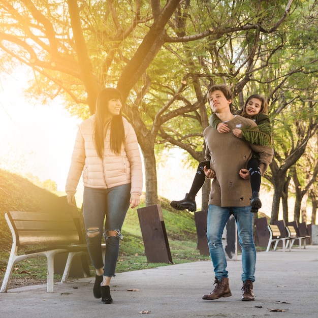 Man giving piggy back to daughter walking in park with his wife