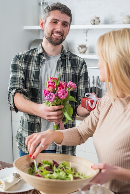 Man giving present and flowers to woman with bowl in kitchen