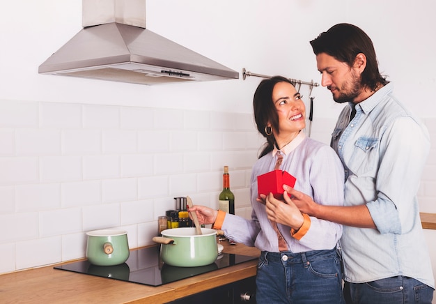 Man giving red gift box to woman in kitchen