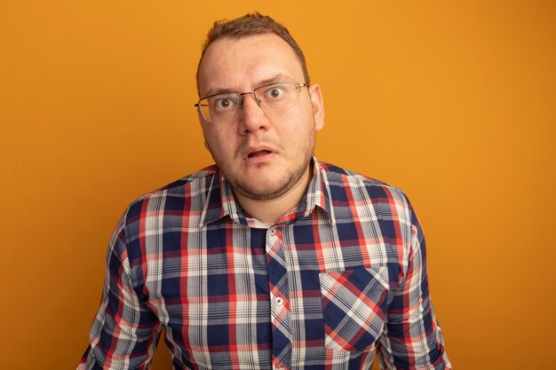 Man in glasses and checked shirt  being confused and worried standing over orange wall