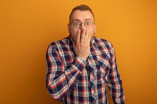 Man in glasses and checked shirt  being shocked covering mouth with hand standing over orange wall