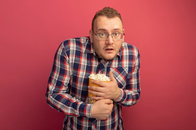 Man in glasses and checked shirt holding bucket with popcorn  confused and scared standing over pink wall