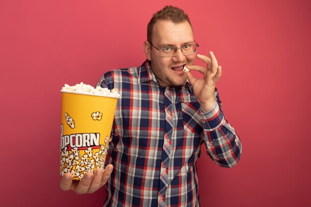 Man in glasses and checked shirt holding bucket with popcorn eating smiling confident standing over pink wall