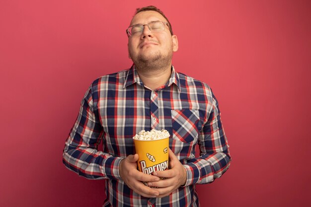 Man in glasses and checked shirt holding bucket with popcorn looking up with closed eyes feeling positive emotions standing over pink wall