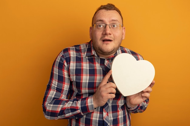 Man in glasses and checked shirt holding cardborad heart looking aside confused standing over orange wall