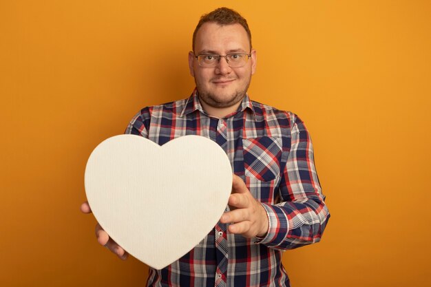 Man in glasses and checked shirt holding cardborad heart  smiling with happy face standing over orange wall