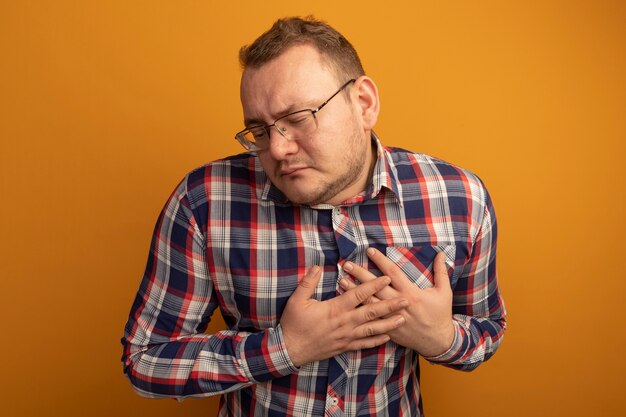 Uomo con gli occhiali e la camicia a quadri tenendo le mani sul petto sentendosi grato in piedi oltre il muro arancione