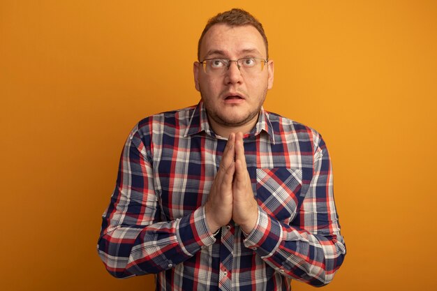 Man in glasses and checked shirt holding hands together like praying looking up with hope expression standing over orange wall