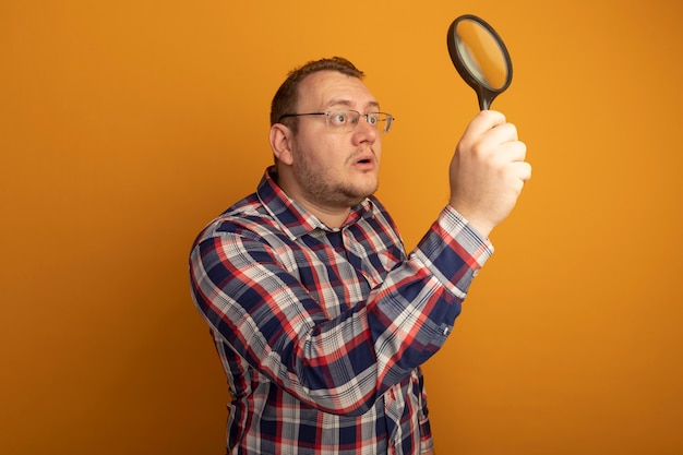 Foto gratuita l'uomo con gli occhiali e la camicia a quadri tenendo la lente d'ingrandimento guardandolo incuriosito in piedi sopra il muro arancione