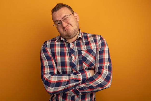 Man in glasses and checked shirt looking aside with skeptic expression making wry mouth standing over orange wall