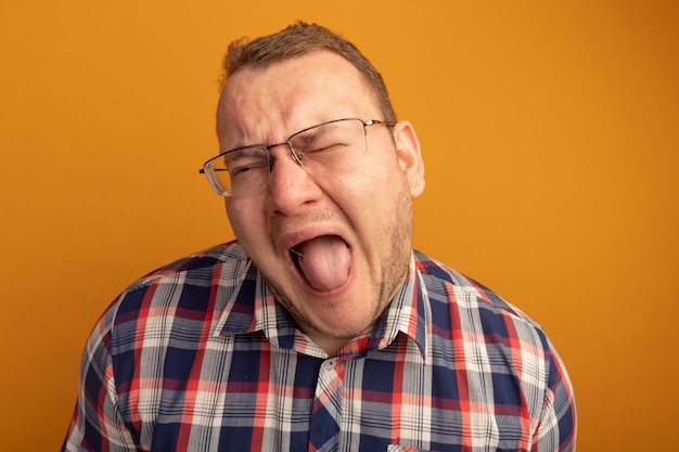 Free photo man in glasses and checked shirt shouting with annoyed expression standing over orange wall