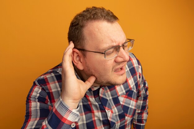 Man in glasses and checked shirt with hand over ear listening to gossips standing over orange wall