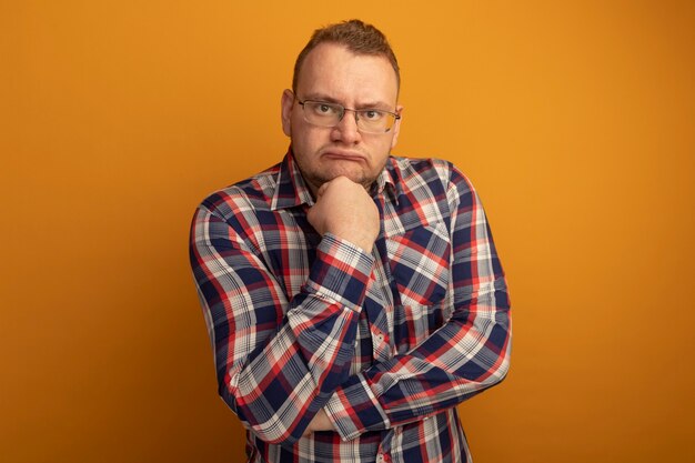 Man in glasses and checked shirt  with hand on his chin thinking with serious face standing over orange wall