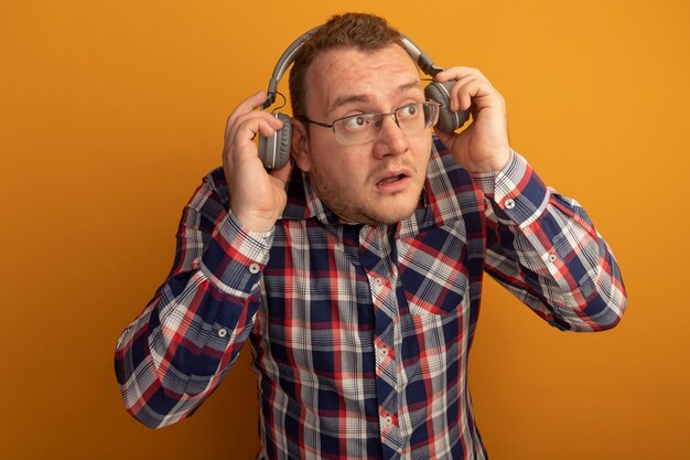 オレンジ色の壁の上に立って混乱している脇に見えるヘッドフォンと眼鏡とチェックシャツの男