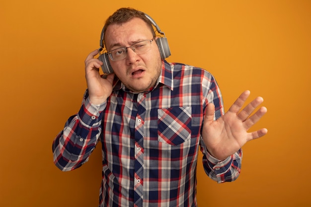 Man in glasses and checked shirt with headphones looking aside confused with raised arm standing over orange wall