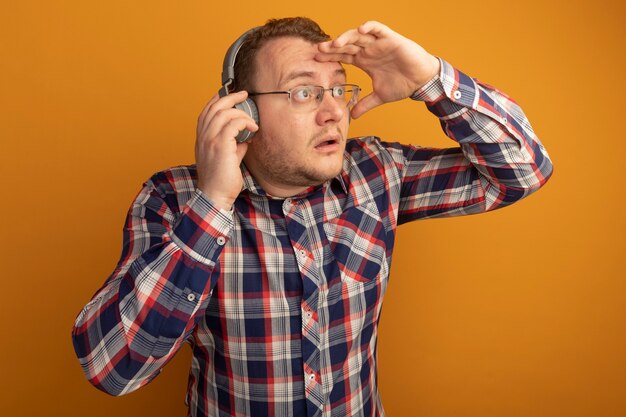 Мужчина в очках и клетчатой рубашке с наушниками смотрит вдаль, подняв руку над головой, стоит над оранжевой стеной