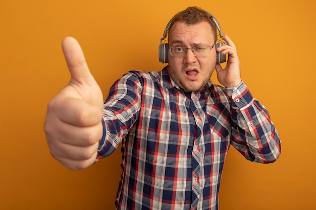 오렌지 벽 위에 서있는 자신감있는 표정으로 엄지 손가락을 보여주는 헤드폰으로 안경과 체크 셔츠를 입은 남자