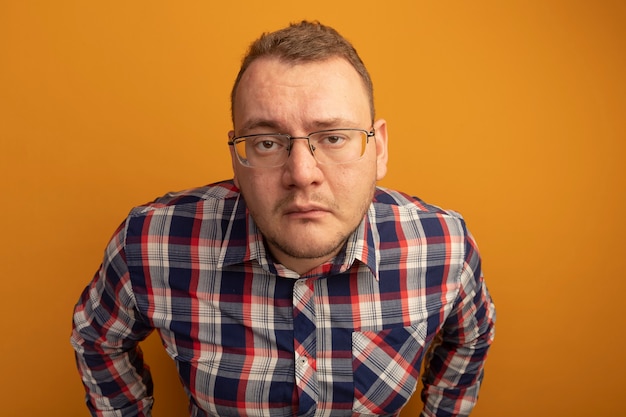 Человек в очках и клетчатой рубашке с грустным выражением лица стоит над оранжевой стеной