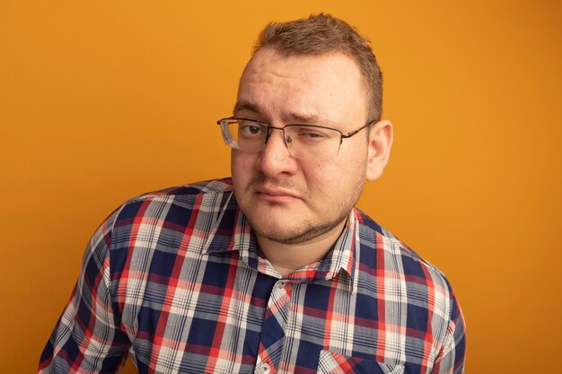 Man in glasses and checked shirt  with skeptic expression standing over orange wall