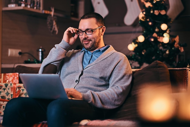Мужчина в очках, одетый в теплый свитер, работает на ноутбуке на Рождество.