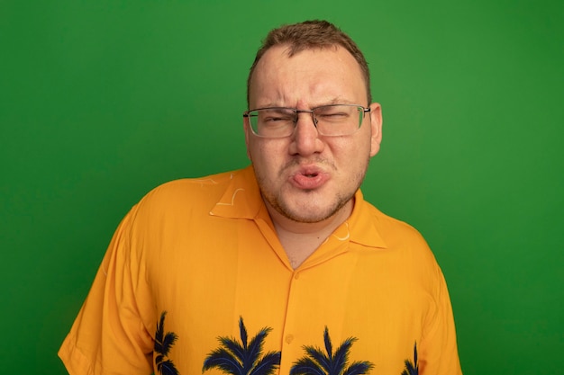 Man in glasses and orange shirt  being displeased frowning standing over green wall