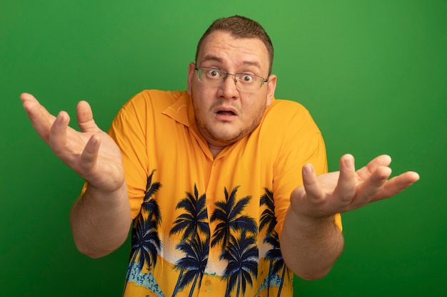 Free photo man in glasses and orange shirt  confused with raised arms having no answer standing over green wall