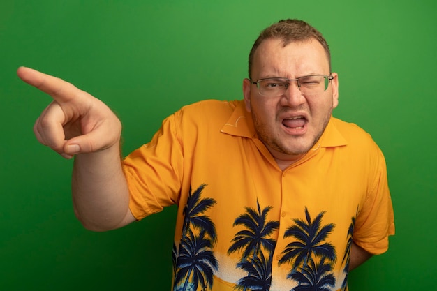 Free photo man in glasses and orange shirt  displeased pointing with finger at something standing over green wall