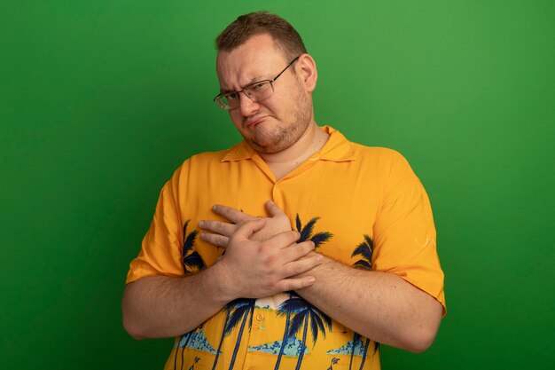 Man in glasses and orange shirt  making wry mouth having disappointed expression standing over green wall