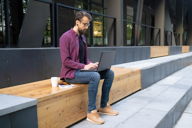 Uomo in vetri che si siedono sul banco di legno e che scrivono sul computer portatile