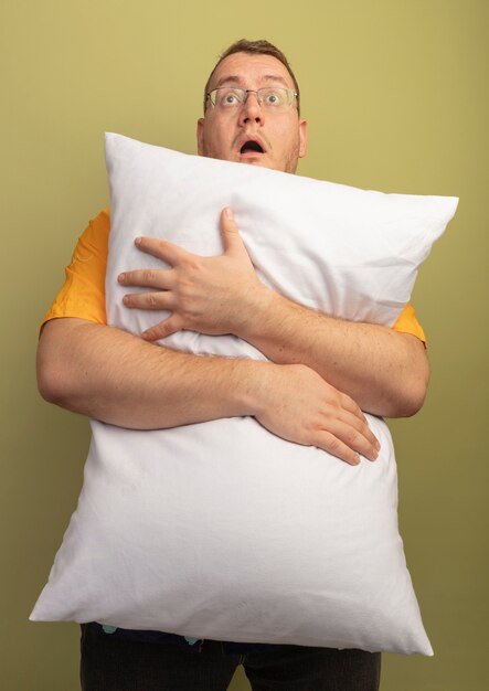 Man in glasses wearing orange shirt hugging pillow looking up surprised and amazed standing over light wall