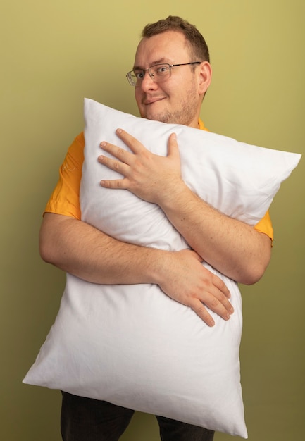 Foto gratuita uomo con gli occhiali che indossa la maglietta arancione che abbraccia il cuscino sorridendo maliziosamente in piedi sopra il muro di luce