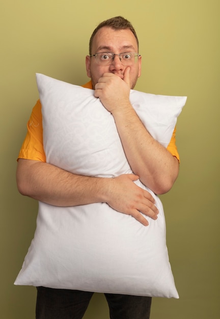 Man in glasses wearing orange shirt hugging pillow  surprised covering mouth with hand standing over light wall