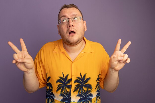 Мужчина в очках в оранжевой рубашке смотрит вверх, показывая смущенный v-знак, стоящий над фиолетовой стеной
