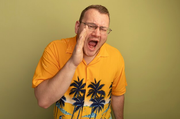Man in glasses wearing orange shirt shouting or calling with hand near mouth standing over light wall