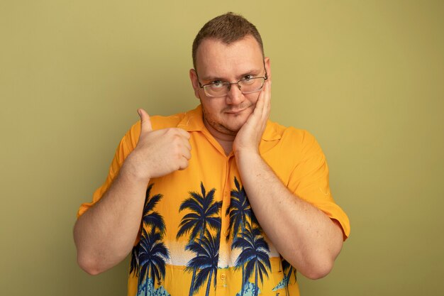 Man in glasses wearing orange shirt  showing thumbs up smiling standing over light wall