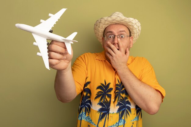 Man in glasses wearing orange shirt in summer hat holding toy airplane  being shocked covering mouth with hand standing over green wall