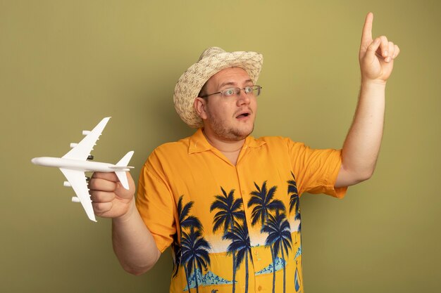 Man in glasses wearing orange shirt in summer hat holding toy airplane looking aside showing index finger surprised and happy standing over green wall