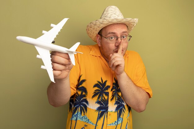 Man in glasses wearing orange shirt in summer hat holding toy airplane making silence gesture with finger on lips standing over green wall