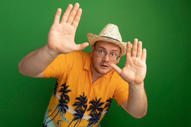 오렌지 셔츠와 여름 모자를 쓰고 손으로 방어 제스처를 만드는 안경에 남자가 녹색 벽 위에 서 무서워하는