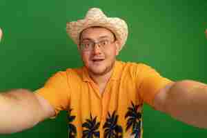 Free photo man in glasses wearing orange shirt and summer hat  with smile on face standing over green wall