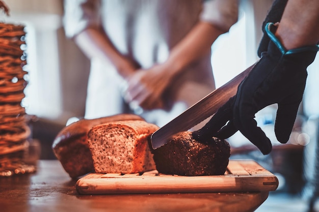 Man in gloves is slicing tasty loaf for lunch at small artisan caffeteria.