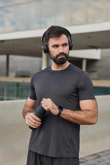Человек занимается спортом делает перерыв слушает мотивационную музыку в беспроводных наушниках освежает водой носит повседневную футболку позирует на размытых упражнениях на открытом воздухе