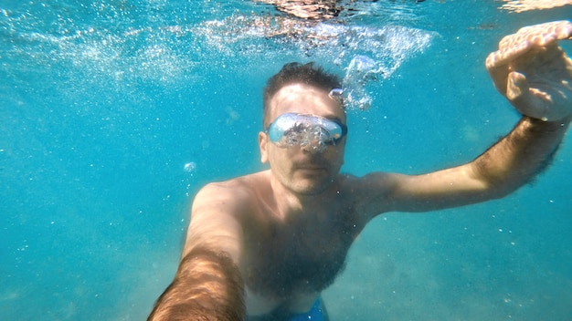 Man in goggles swimming under the blue and transparent water of the Mediterranean sea. Holding the camera