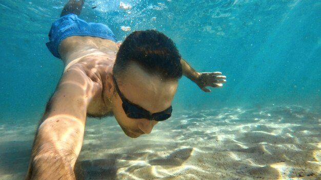 Man in goggles swimming under the blue and transparent water of the Mediterranean sea. Holding the camera