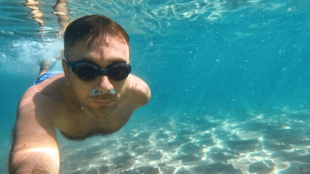 Man in goggles swimming under the blue and transparent water of the Mediterranean sea. Holding the camera