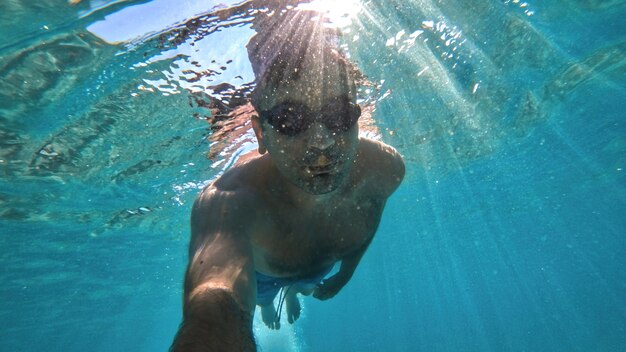 Man in goggles swimming under the blue and transparent water of the Mediterranean sea. Holding the camera