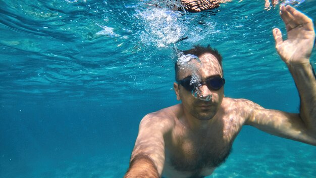 Man in goggles swimming under the blue and transparent water of the Mediterranean sea. Holding the camera