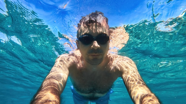 Man in goggles swimming under the blue and transparent water of the Mediterranean sea. Holding the camera