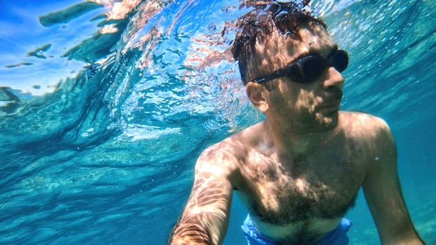 Man in goggles swimming under the blue and transparent water of the Mediterranean sea. Holding the camera