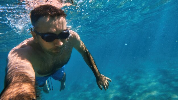 Man in goggles swimming under the blue and transparent water of the Mediterranean sea. Holding the camera